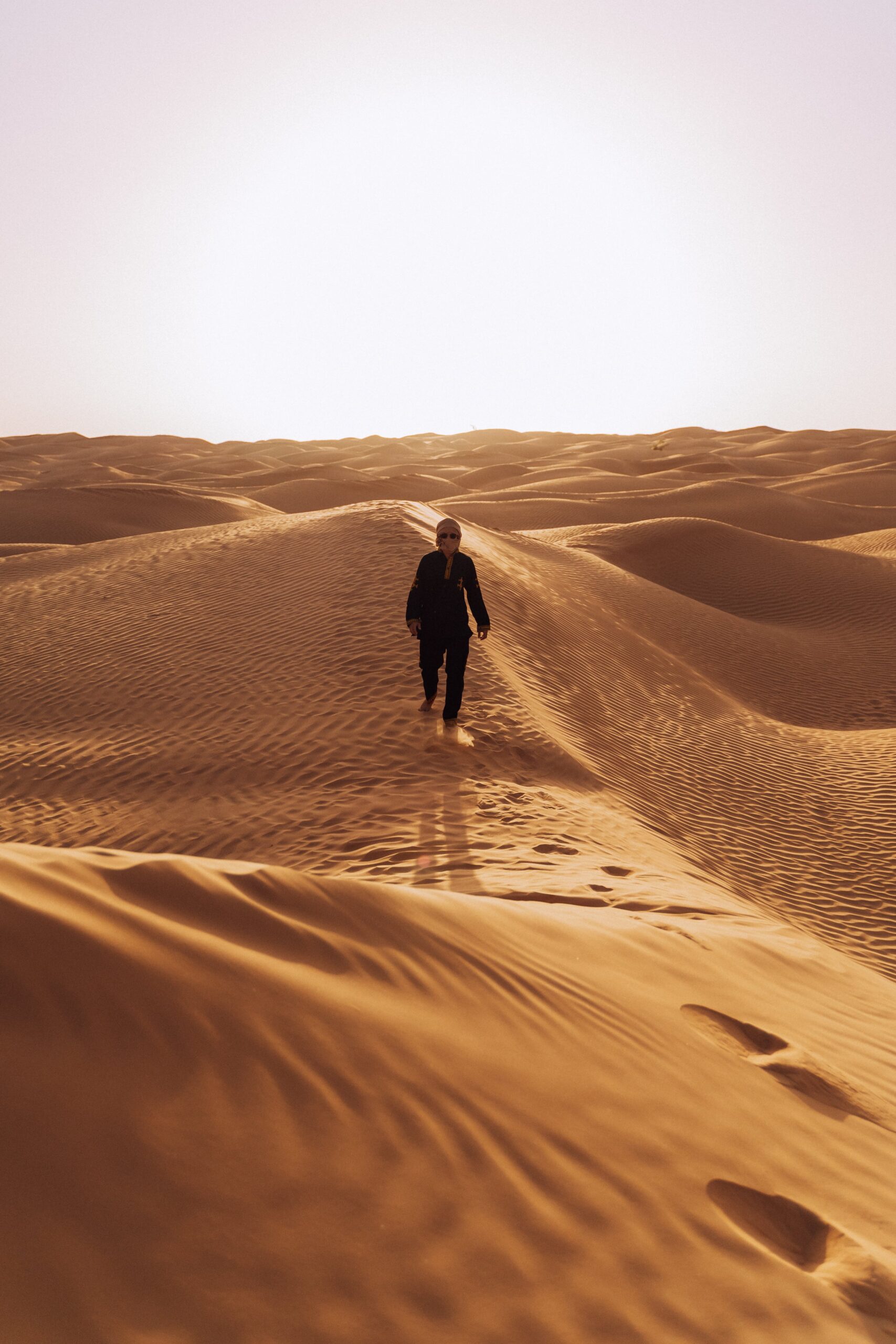 Traveler walking across golden sand dunes in the Sahara Desert, Tunisia, with footprints trailing behind.